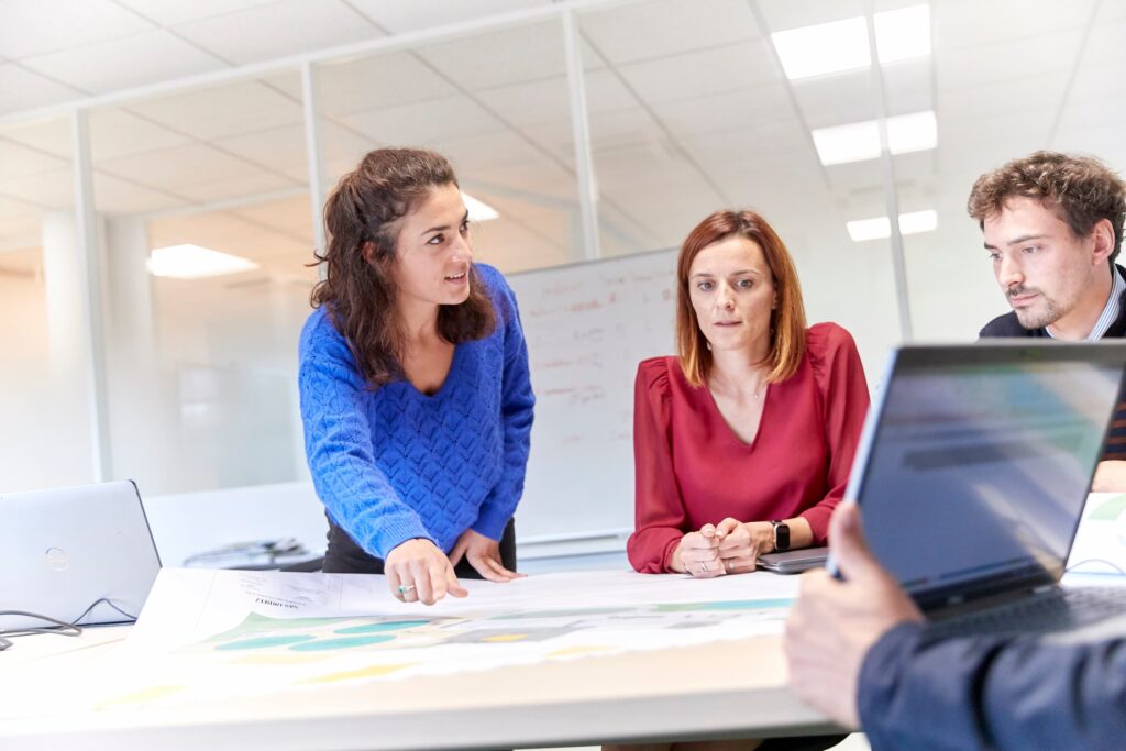 Réunion d’équipe au bureau autour de plans, avec trois collaborateurs devant des ordinateurs.