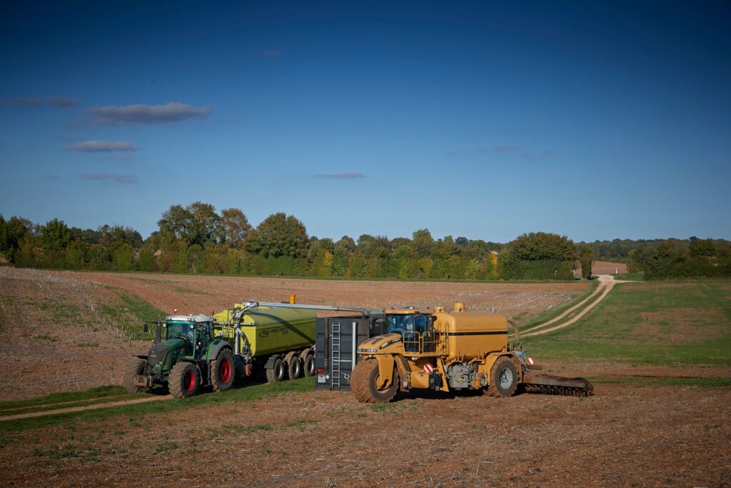 Tracteurs et épandeur agricole en train d’épandre du lisier sur un champ, sous un ciel bleu.